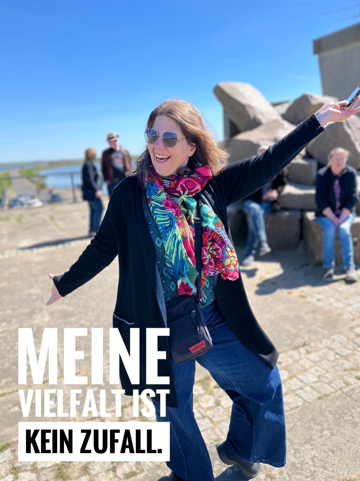 Lachende Frau mit Sonnenbrille am Wasser mit dem Statement „Meine Vielfalt ist kein Zufall“ für Persönlichkeit, Diversität und Entwicklungslust.
